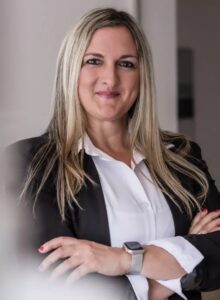 Eine lächelnde Frau mit blondem Haar in schwarzem Blazer und weißem Hemd, trägt eine Armbanduhr.
